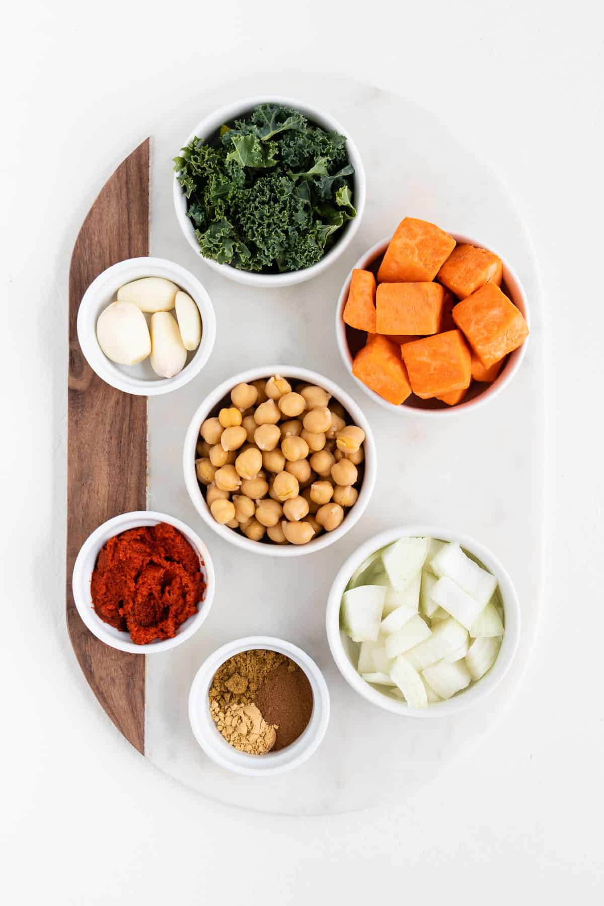 bowls filled with cubed potatoes, garbanzo beans, curry paste, spices, onion, greens, and garlic on a white marble board