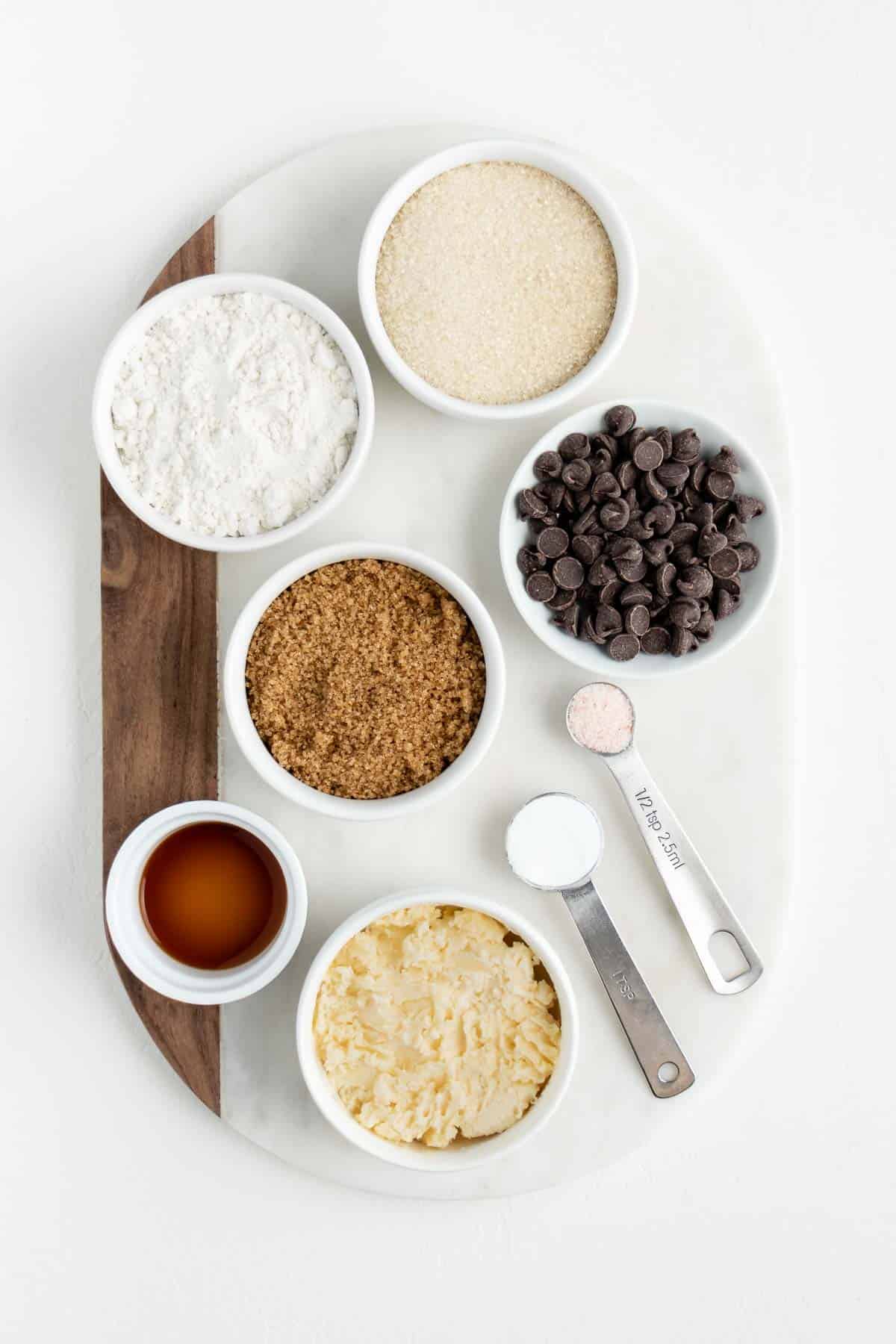 white bowls filled with flour, sugar, vegan butter, brown sugar, vanilla extract, baking powder, and salt on a white marble board