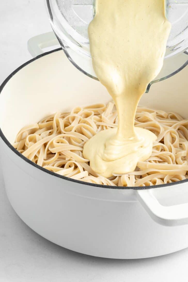 pouring vegan alfredo sauce over a pot of fettuccine noodles