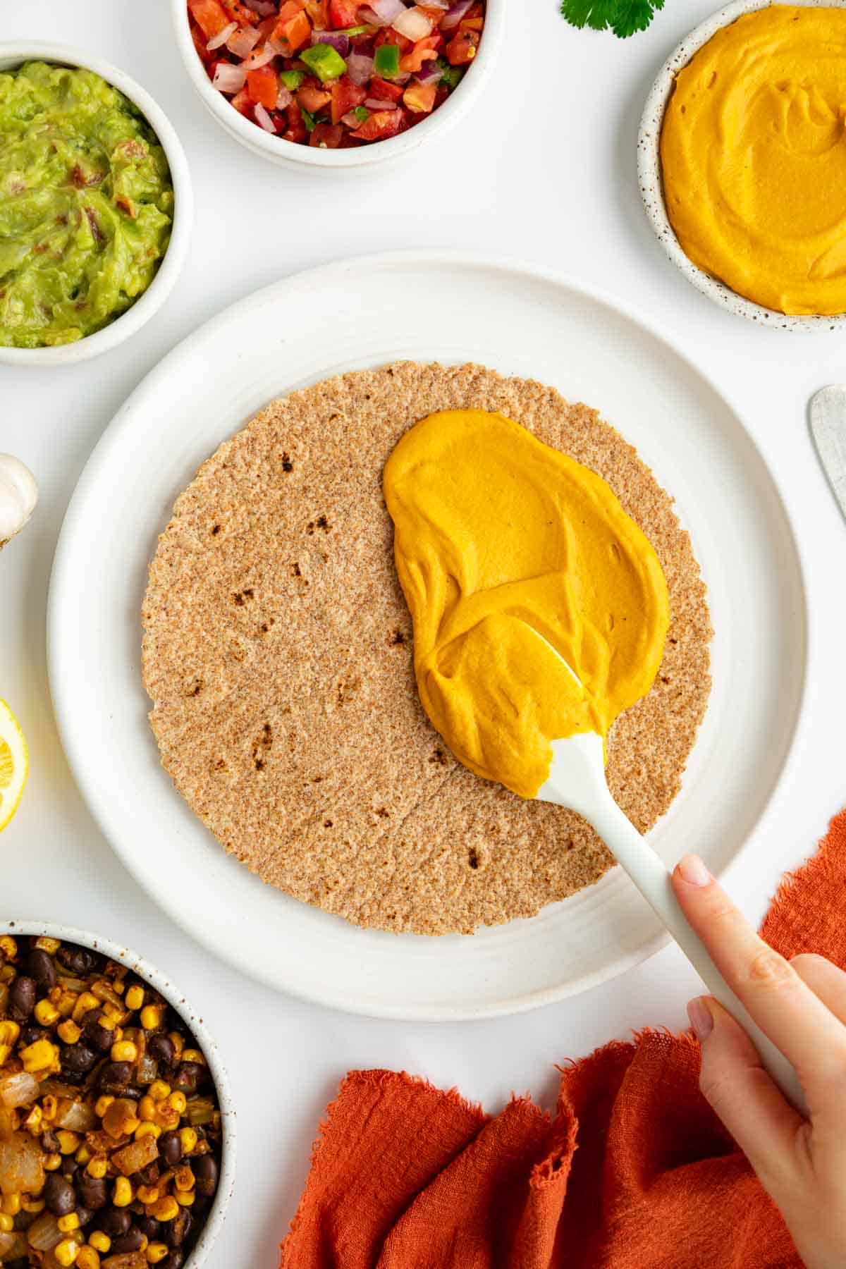 a hand spreading sweet potato cheese on a tortilla, surrounded by bowls of ingredients