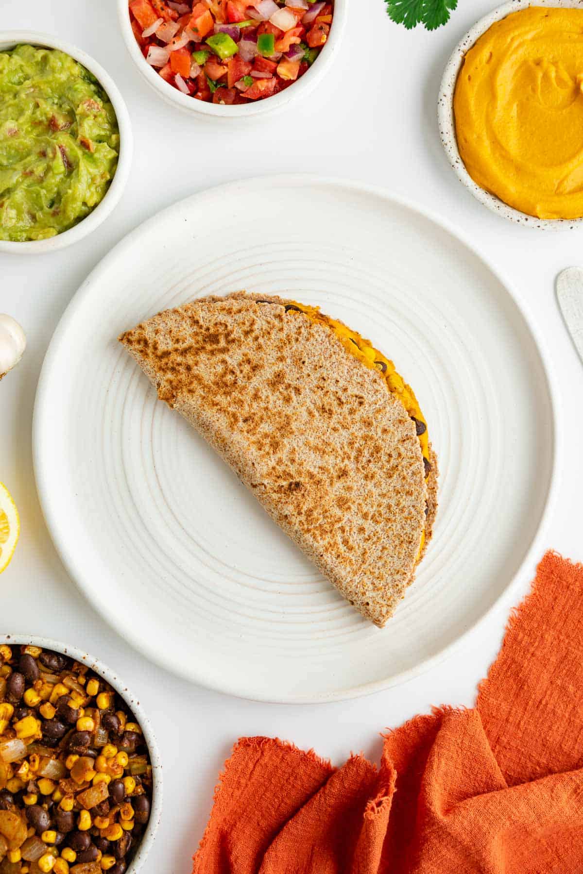 a grilled quesadilla on a white plate surrounded by bowls of ingredients, including guacamole and pico de gallo