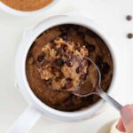 a spoon scooping into a vegan chocolate chip banana mug cake beside a bowl of almond butter