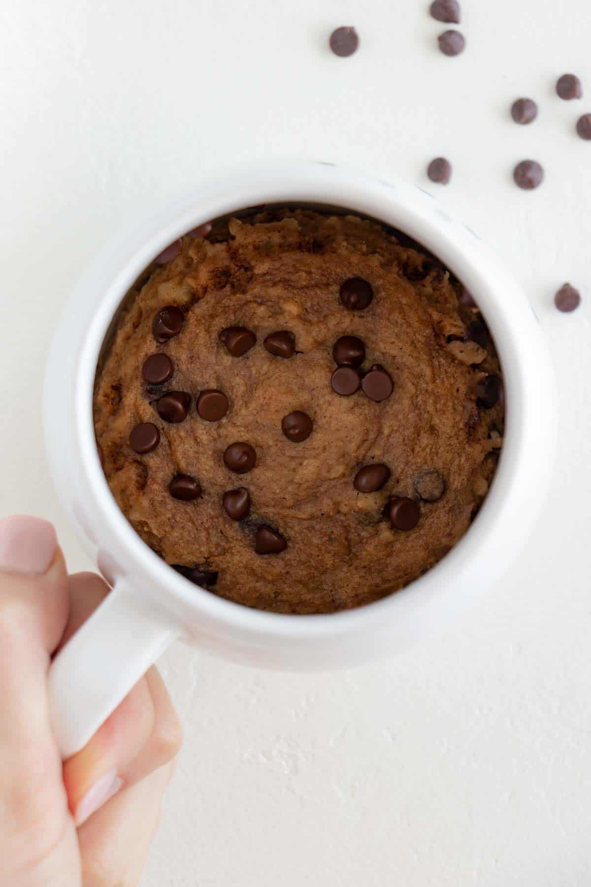 a hand holding a white mug filled with vegan banana bread mug cake topped with chocolate chips inside
