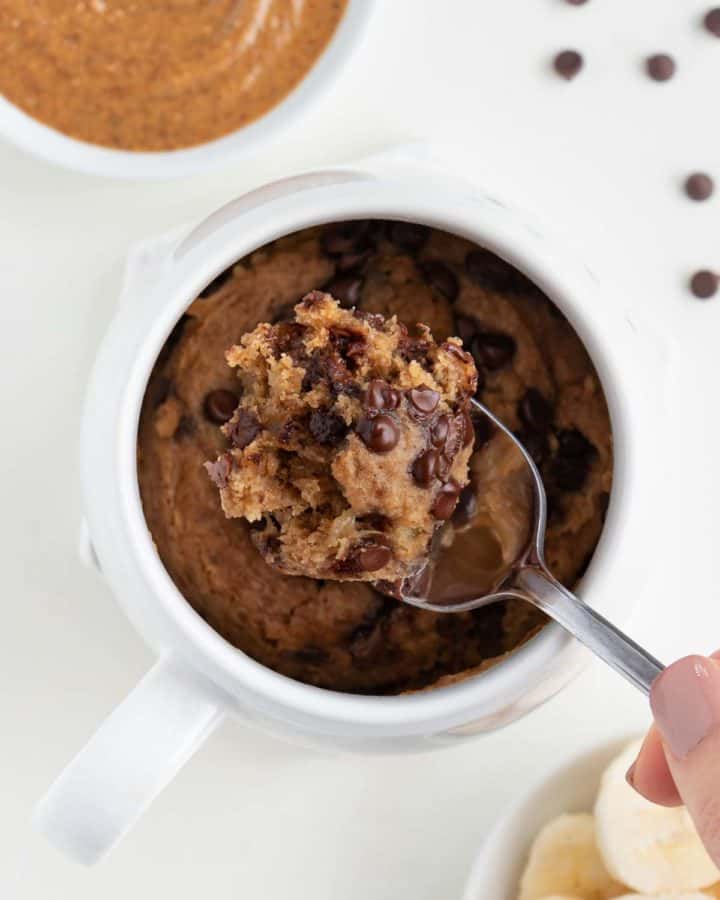 a spoon scooping into a vegan chocolate chip banana mug cake beside a bowl of almond butter