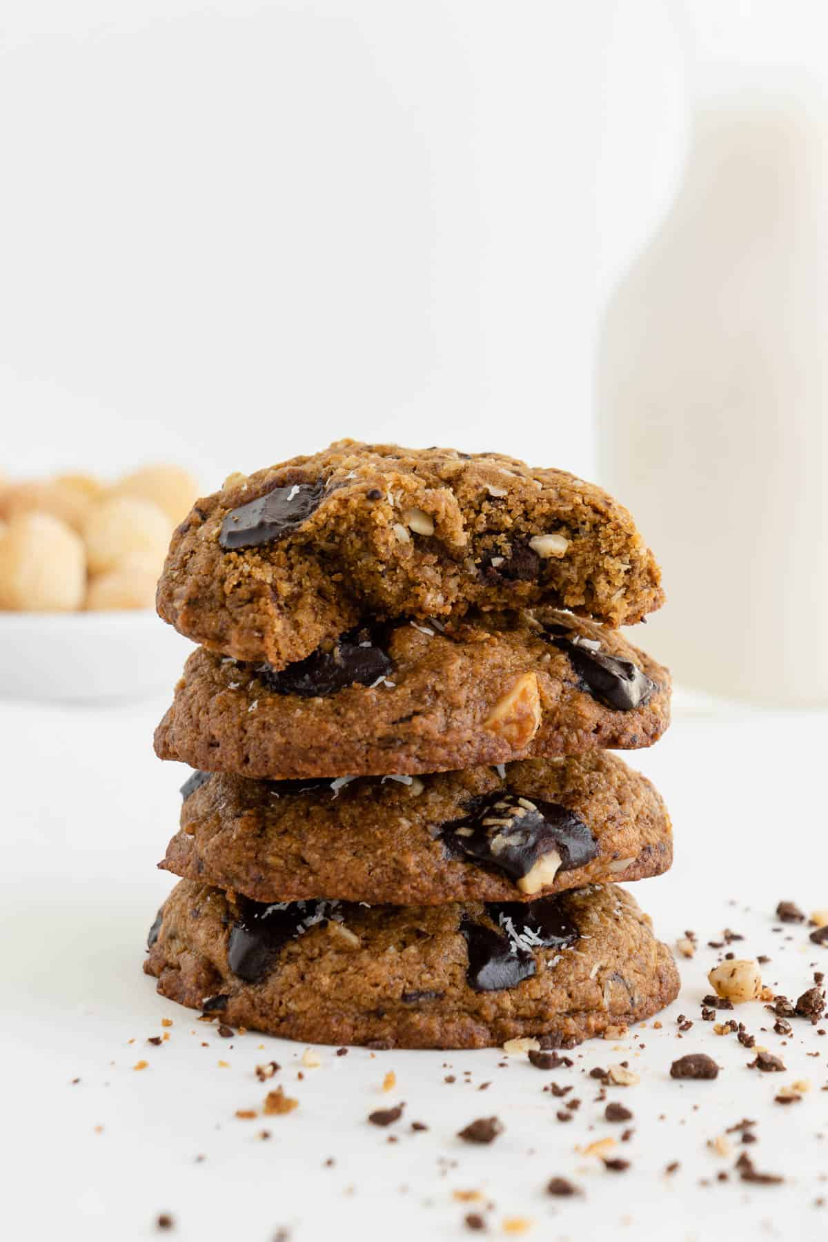 four vegan chocolate chunk hawaiian cookies stacked on top of each other with a glass of almond milk and a bowl of macadamia nuts in the background