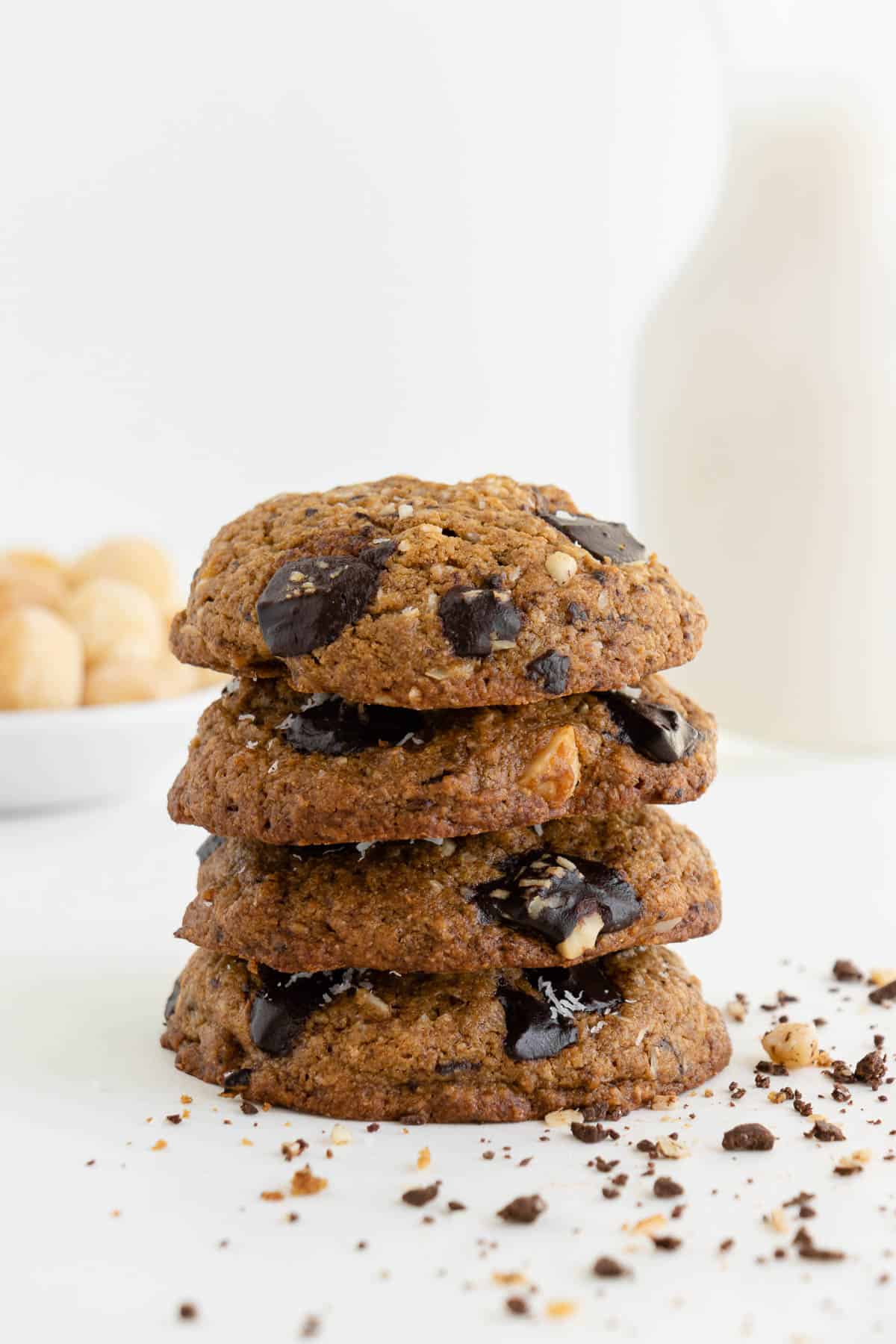 four vegan chocolate chunk hawaiian cookies stacked on top of each other with a glass of almond milk and a bowl of macadamia nuts in the background