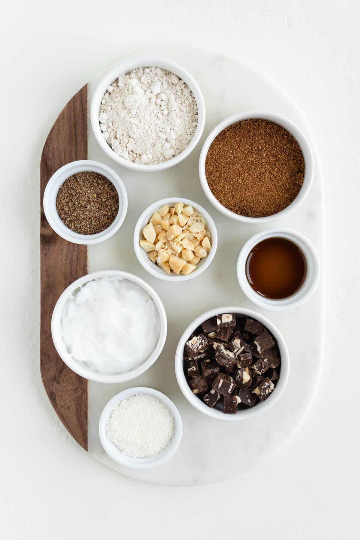 a marble board topped with bowls filled with flour, coconut sugar, shredded coconut, macadamia nuts, chocolate, coconut oil, and ground flaxseed