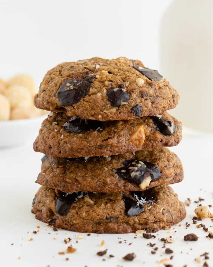 four vegan chocolate chunk hawaiian cookies stacked on top of each other with a glass of almond milk and a bowl of macadamia nuts in the background
