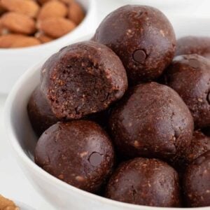 no bake brownie bites stacked inside a white bowl surrounded by walnuts, almonds, and chocolate chips