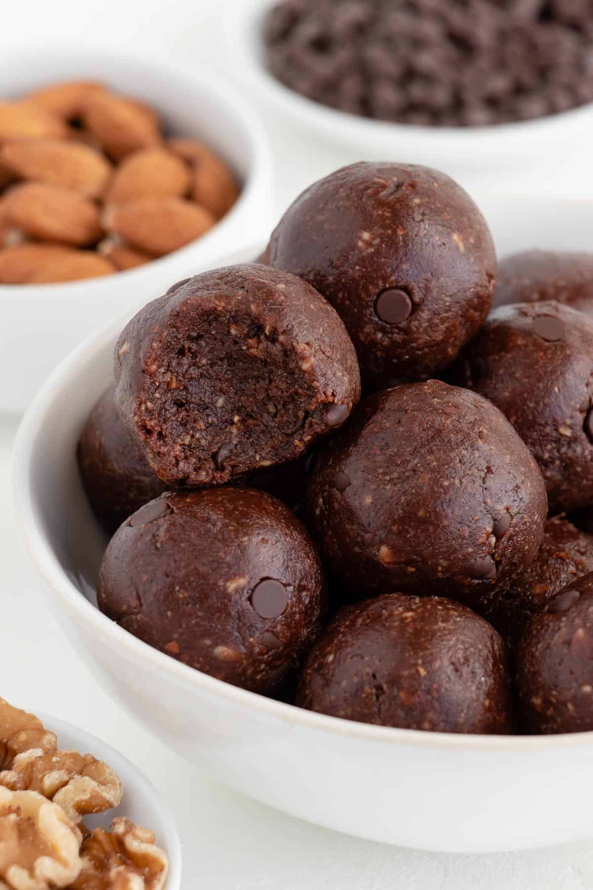 no bake brownie bites stacked inside a white bowl surrounded by walnuts, almonds, and chocolate chips