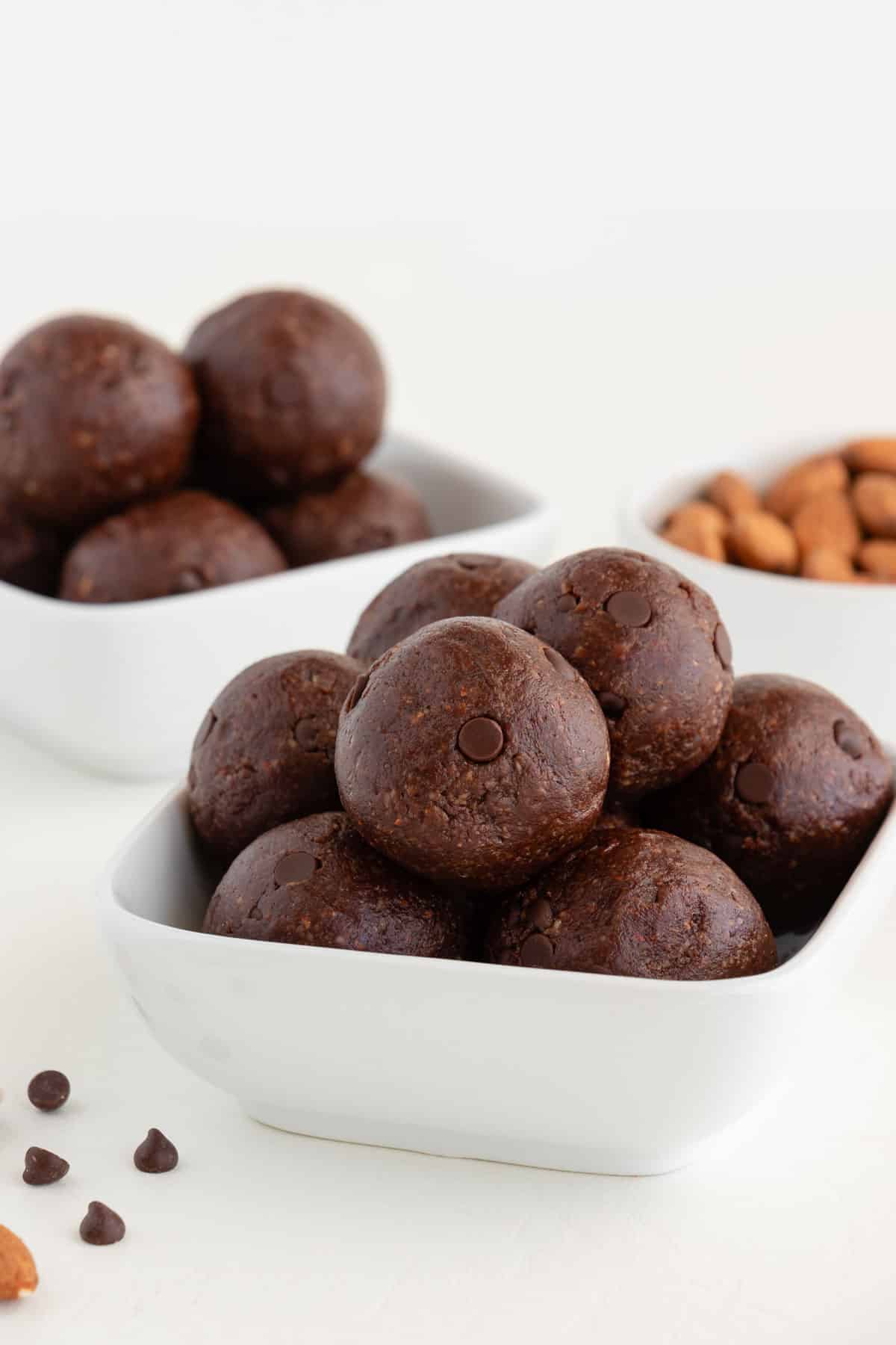 no bake brownie bites inside two white bowls beside chocolate chips and almonds