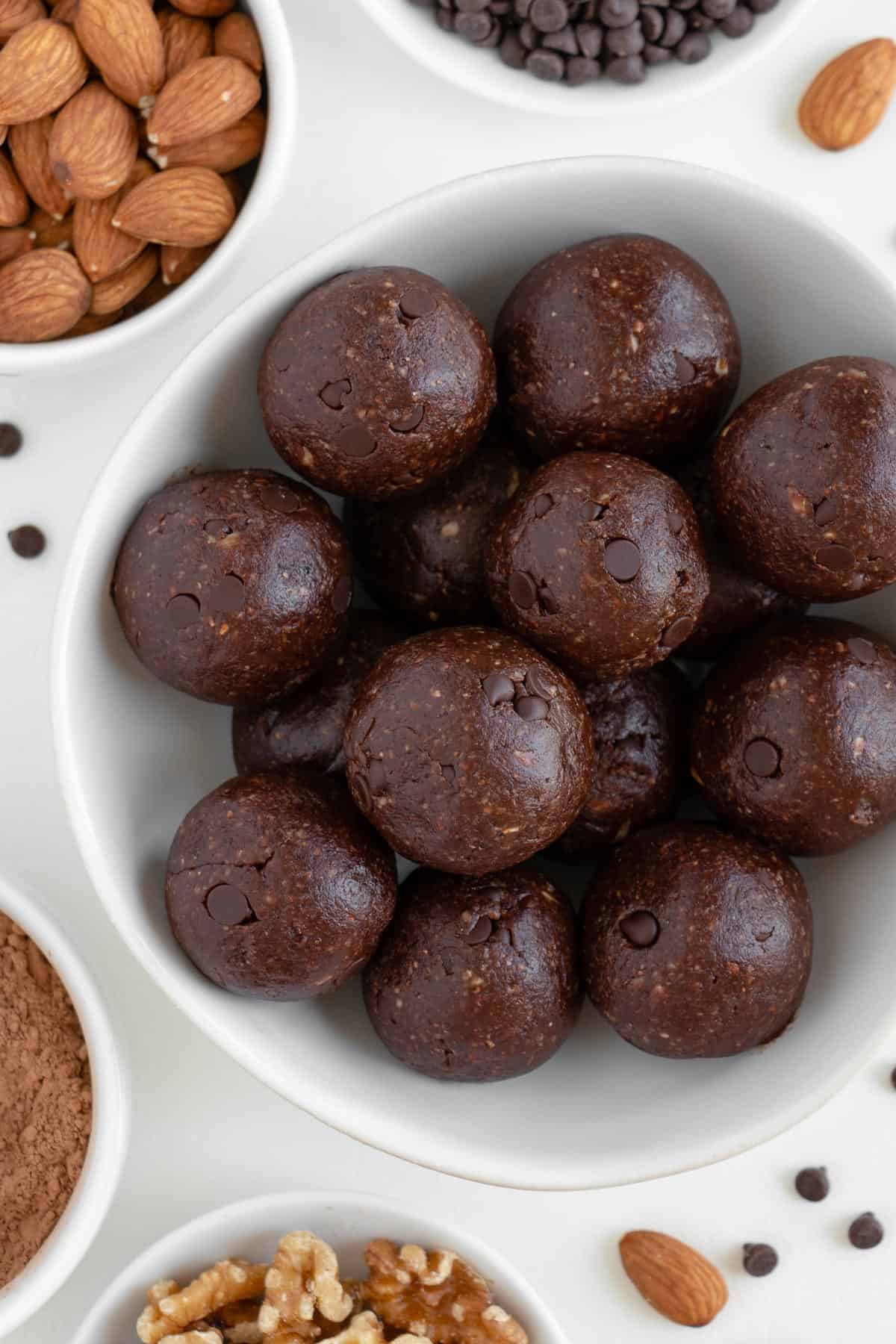 no bake brownie bites in a white bowl surrounded by almonds, chocolate chips, cacao powder, and walnuts