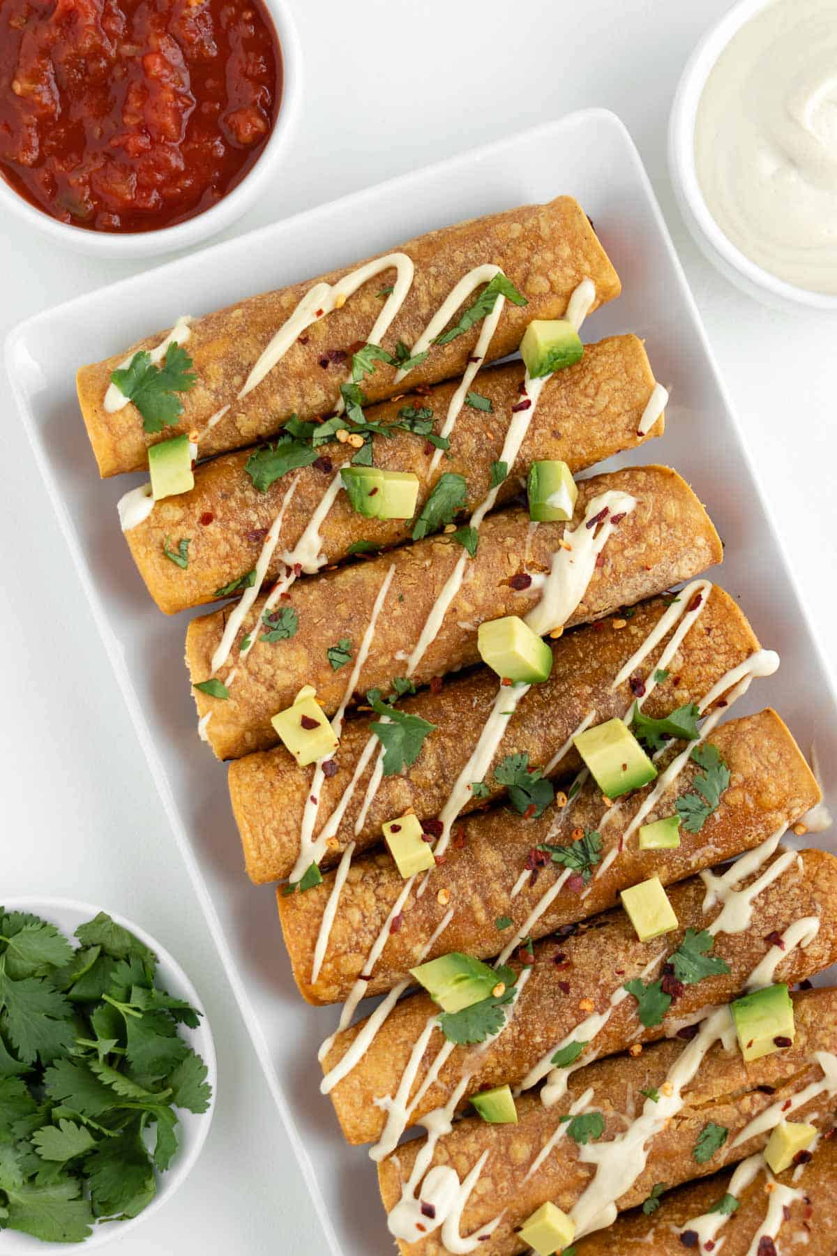 sweet potato black bean taquitos on a white plate surrounded by guacamole, salsa, cilantro, and vegan sour cream