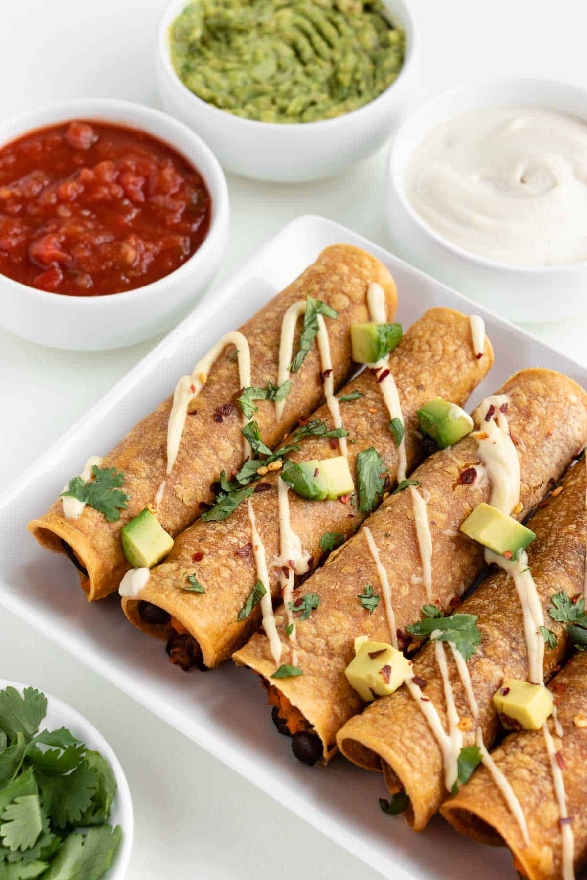 vegan sweet potato and black bean taquitos on a white plate beside salsa, guacamole, and sour cream