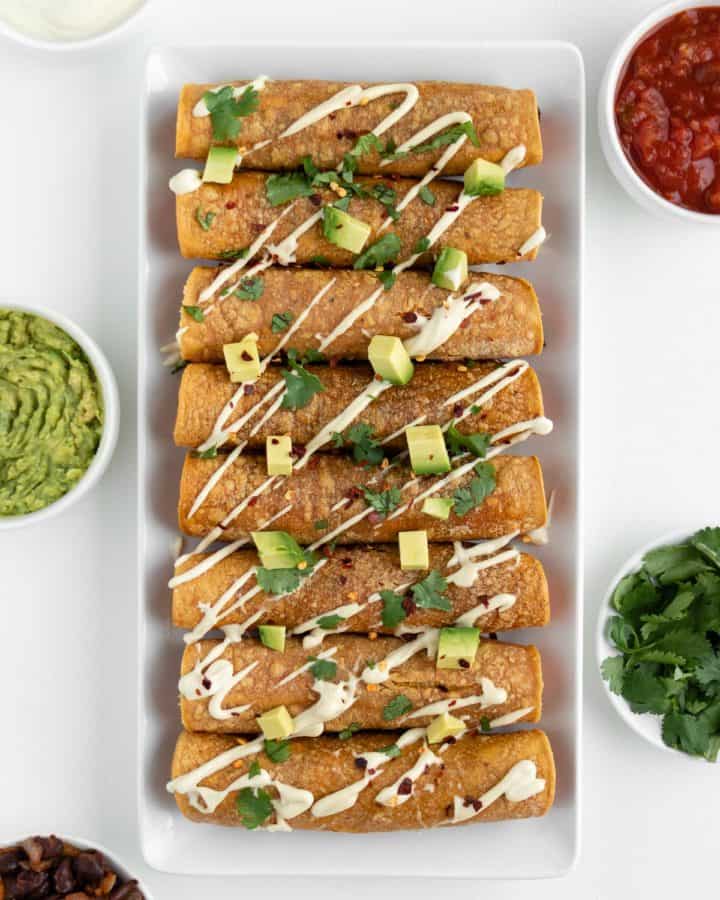 sweet potato black bean taquitos on a white plate surrounded by guacamole, salsa, cilantro, and vegan sour cream