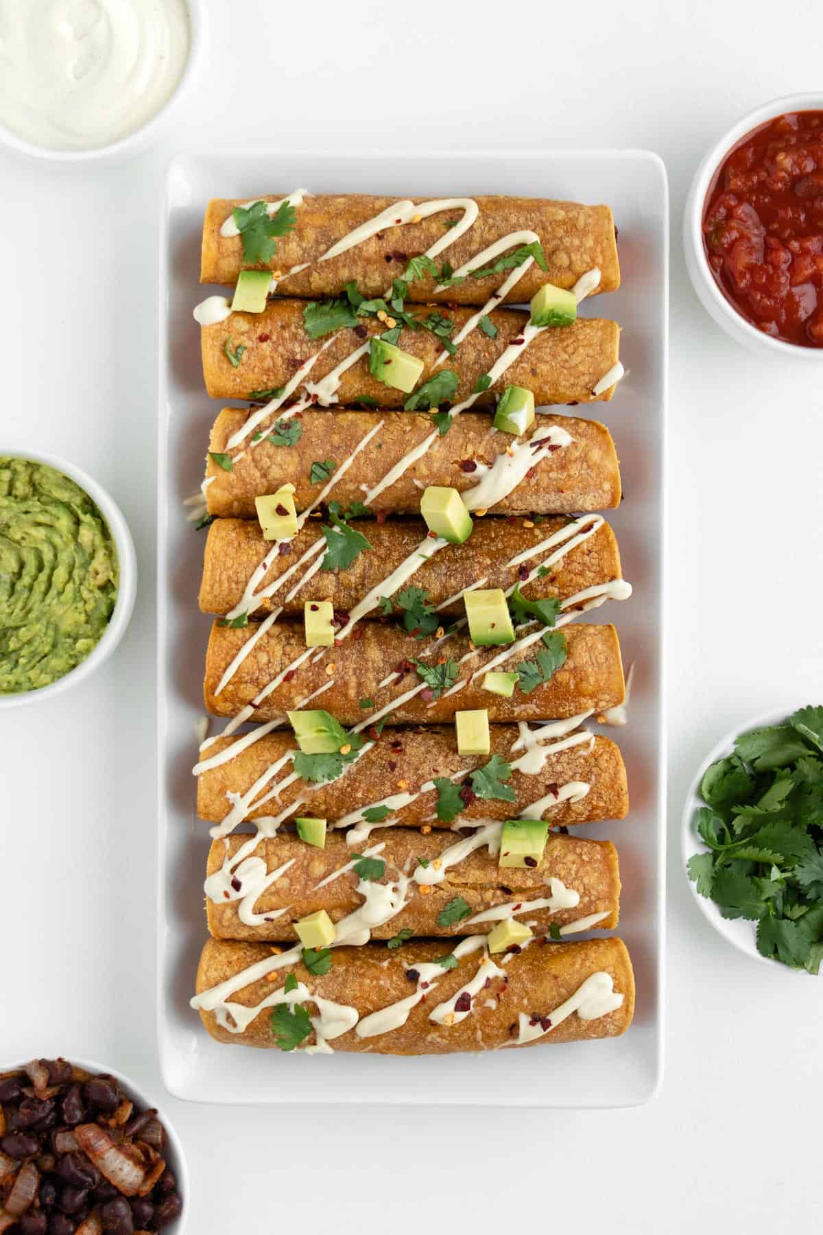 sweet potato black bean taquitos on a white plate surrounded by guacamole, salsa, cilantro, and vegan sour cream
