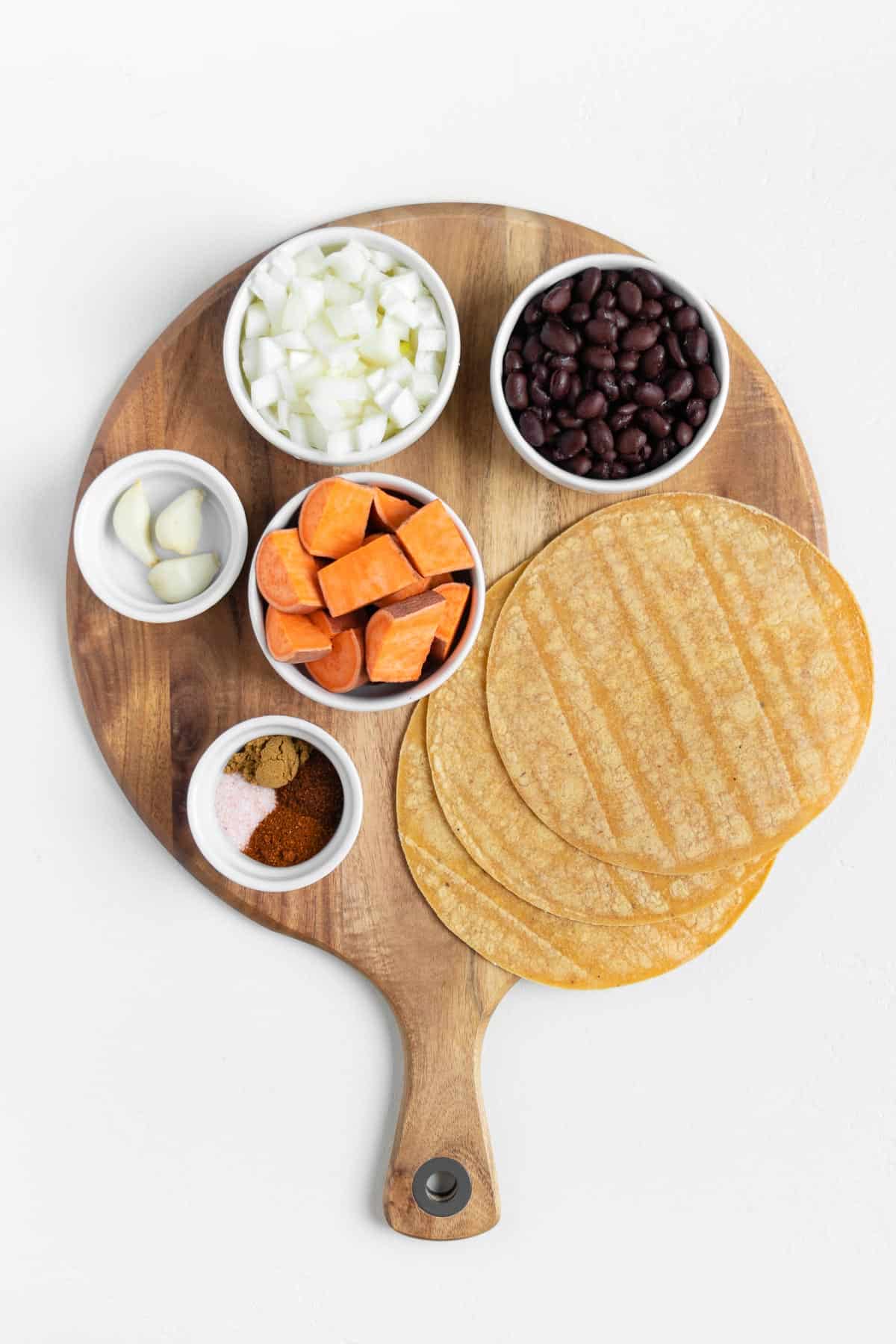 white bowls filled with cubed yams, beans, onion, garlic, spices, and corn tortillas on a round wooden cutting board