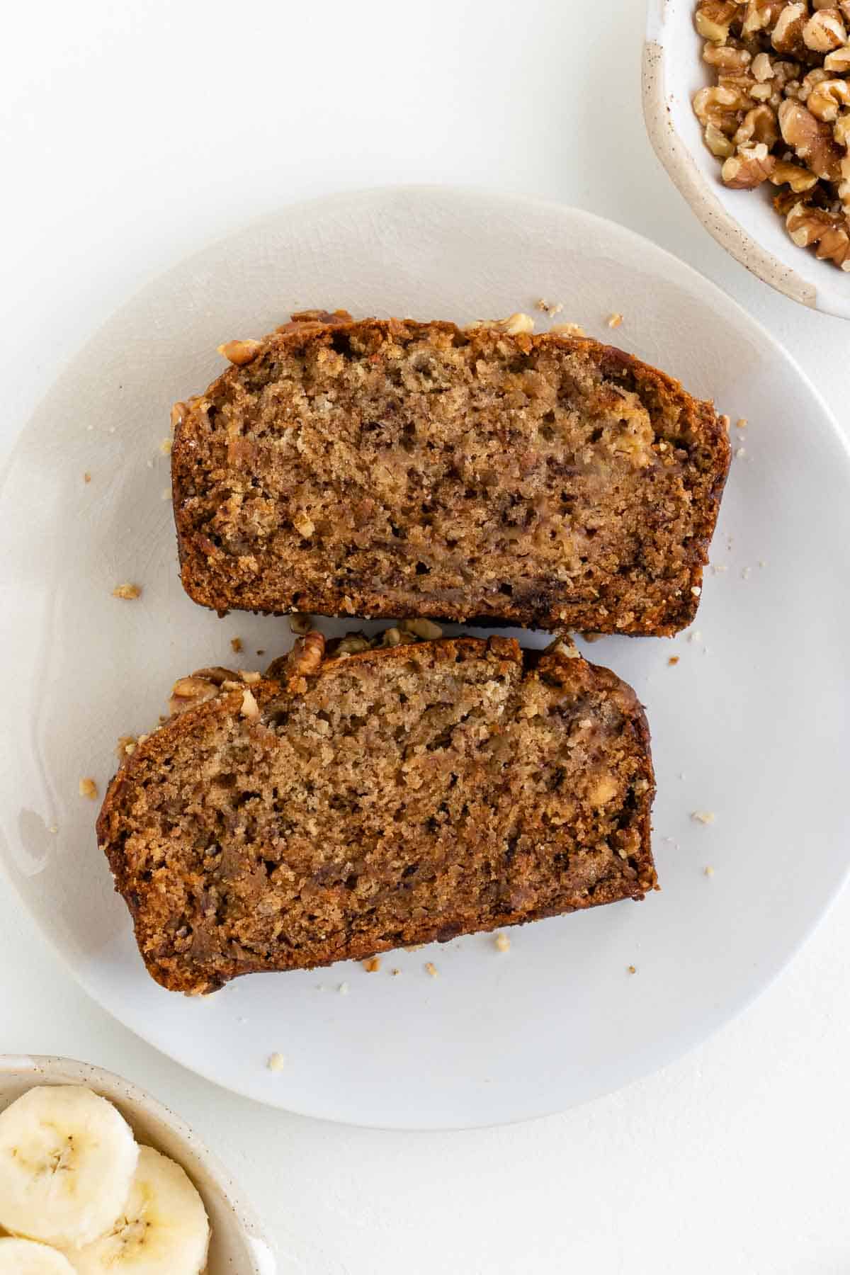 two slices of vegan banana bread on a plate beside a bowl of walnuts and a bowl of sliced banana