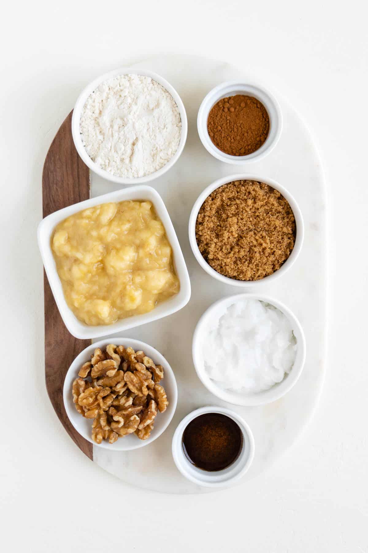 mashed bananas, brown sugar, walnuts, flour, cinnamon, and vanilla extract in white bowls on a marble board