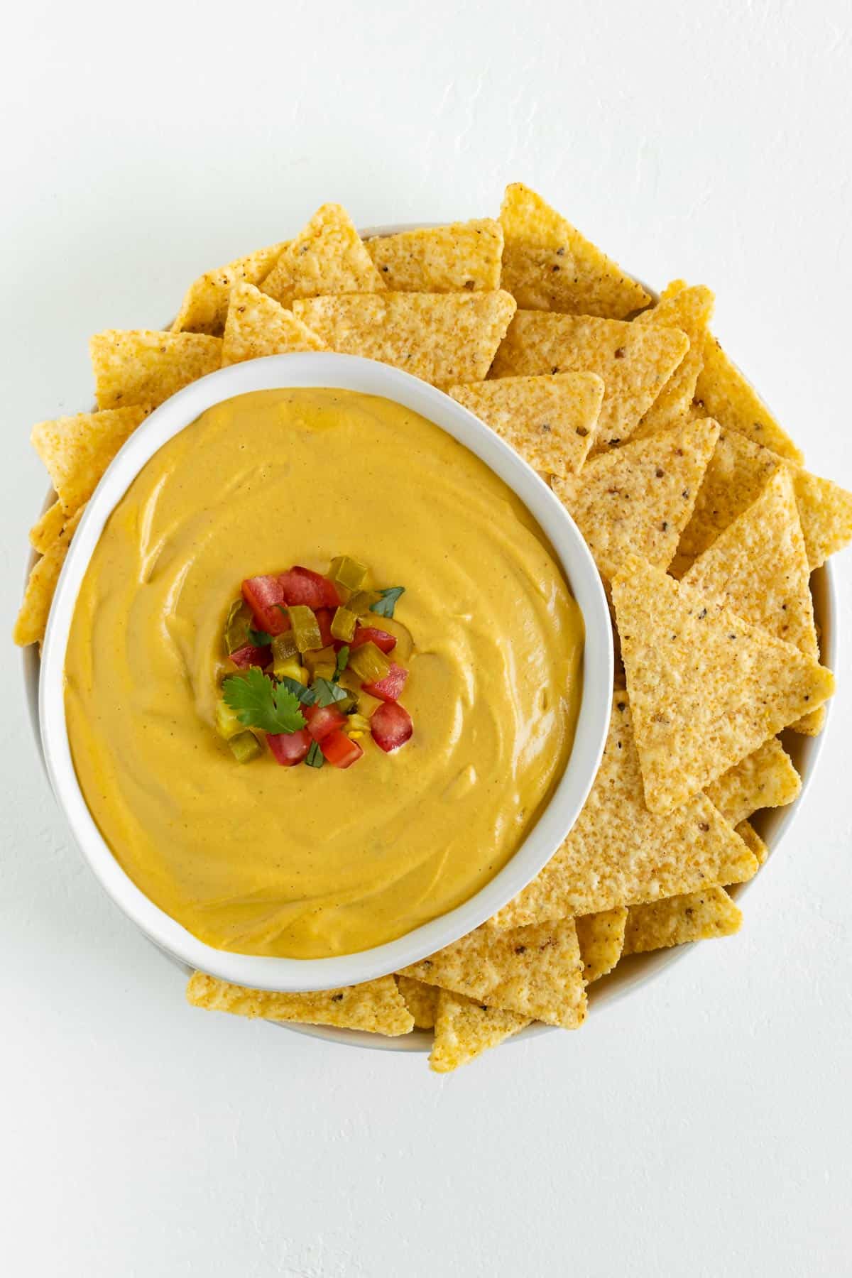 a bowl of vegan nacho cheese garnished with diced jalapeño, tomato, and cilantro beside corn tortilla chips