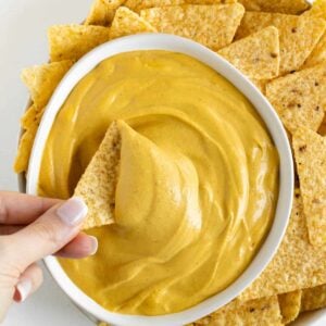 a hand dipping a corn tortilla chip into a bowl of vegan nacho cheese surrounded by more tortilla chips