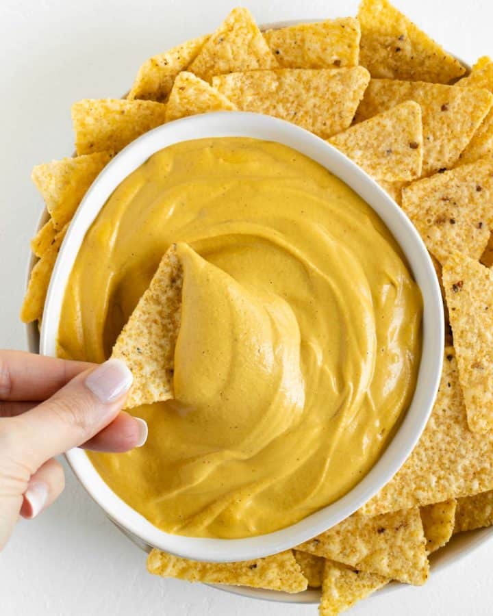 a hand dipping a corn tortilla chip into a bowl of vegan nacho cheese surrounded by more tortilla chips