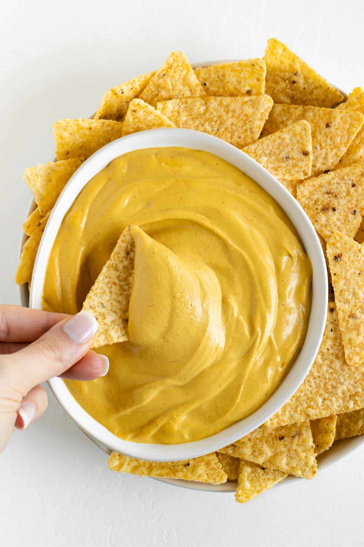 a hand dipping a corn tortilla chip into a bowl of vegan nacho cheese surrounded by more tortilla chips