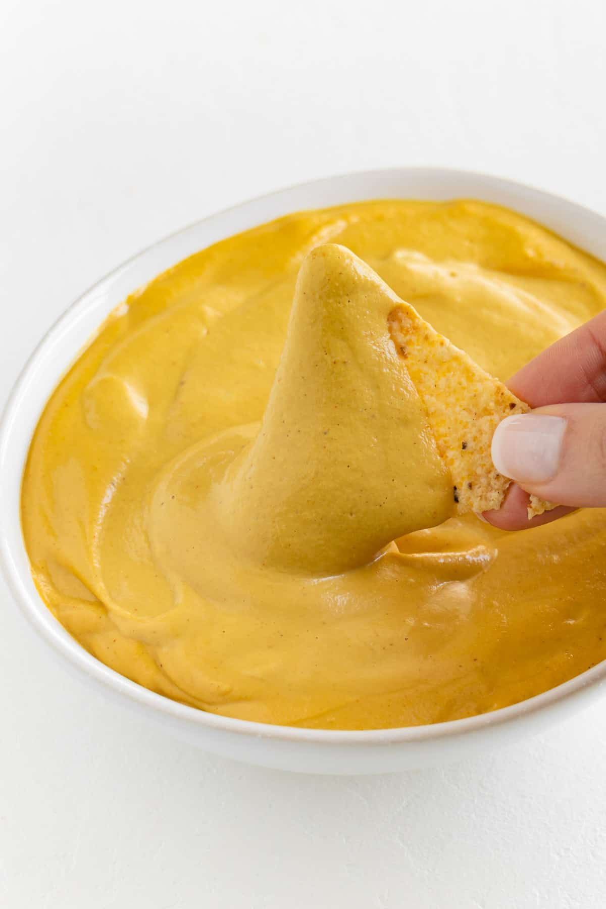 a hand dipping a corn tortilla chip into a bowl of vegan nacho cheese