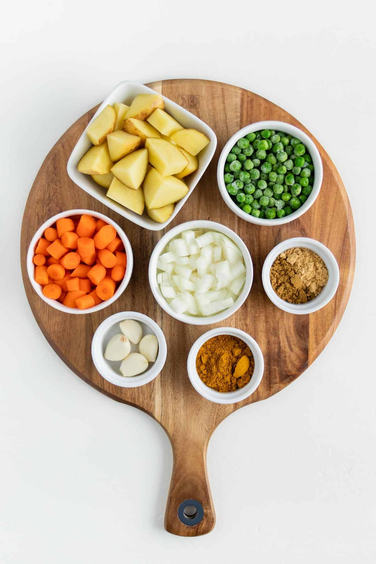 small white bowls filled with potatoes, green peas, carrots, onion, garlic, and spices on a round wooden board