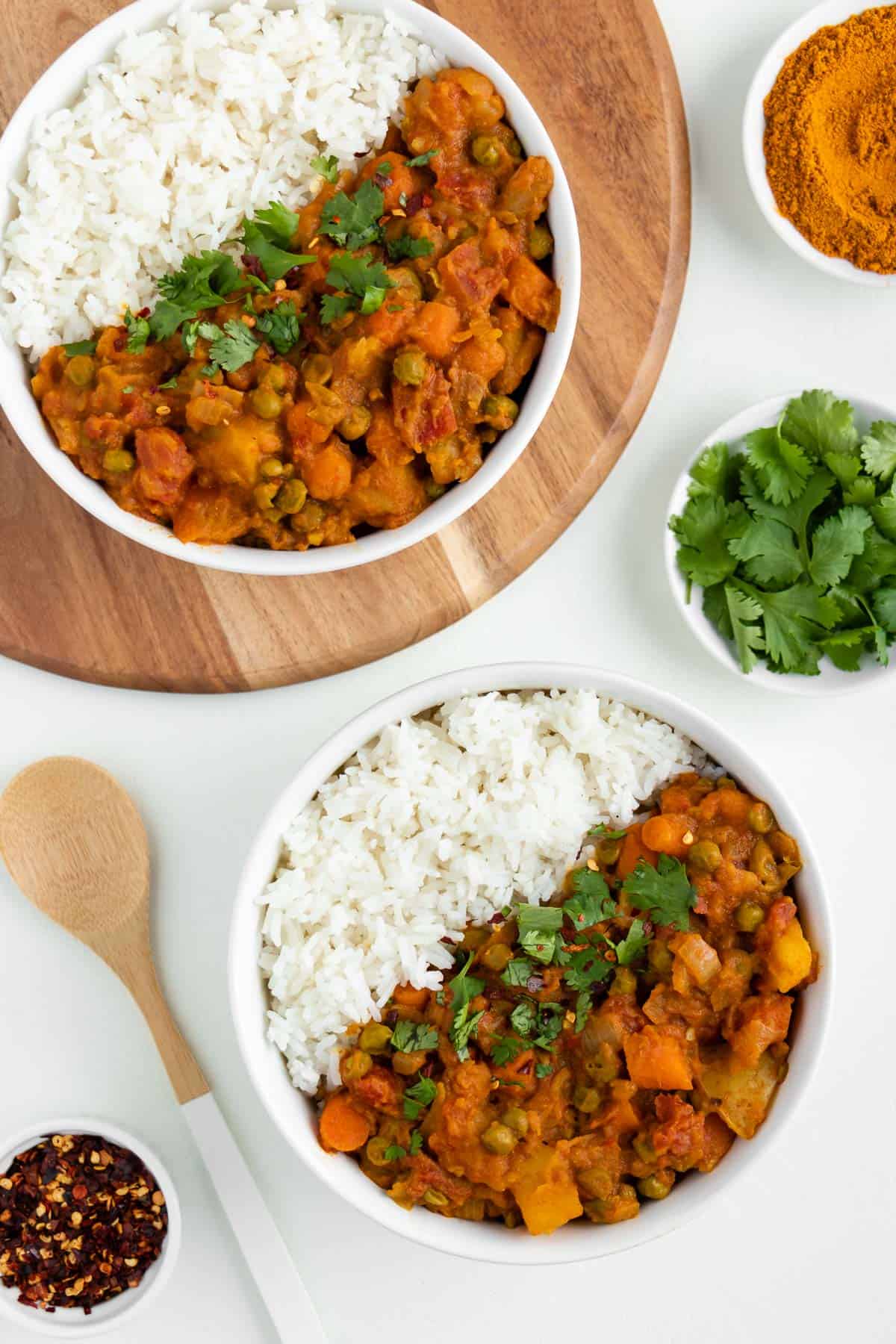 two bowls of vegan potato curry with rice beside a wooden spoon, bowl of red pepper flakes, and bowl of cilantro