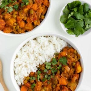 two bowls of vegan potato curry with rice beside a wooden spoon, bowl of red pepper flakes, and bowl of cilantro