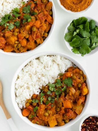 two bowls of vegan potato curry with rice beside a wooden spoon, bowl of red pepper flakes, and bowl of cilantro