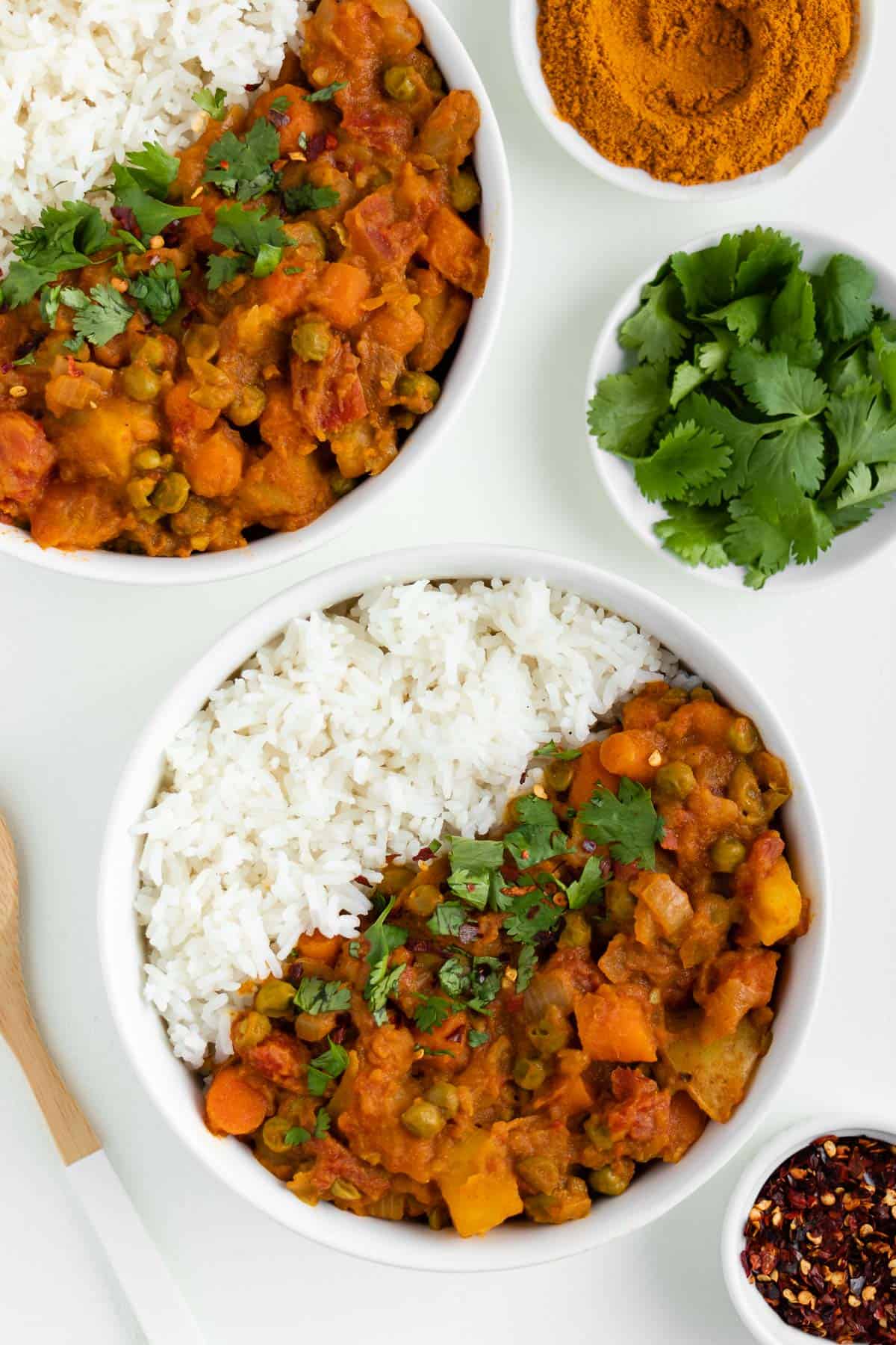 two bowls of vegan potato curry with rice beside a wooden spoon, bowl of red pepper flakes, and bowl of cilantro