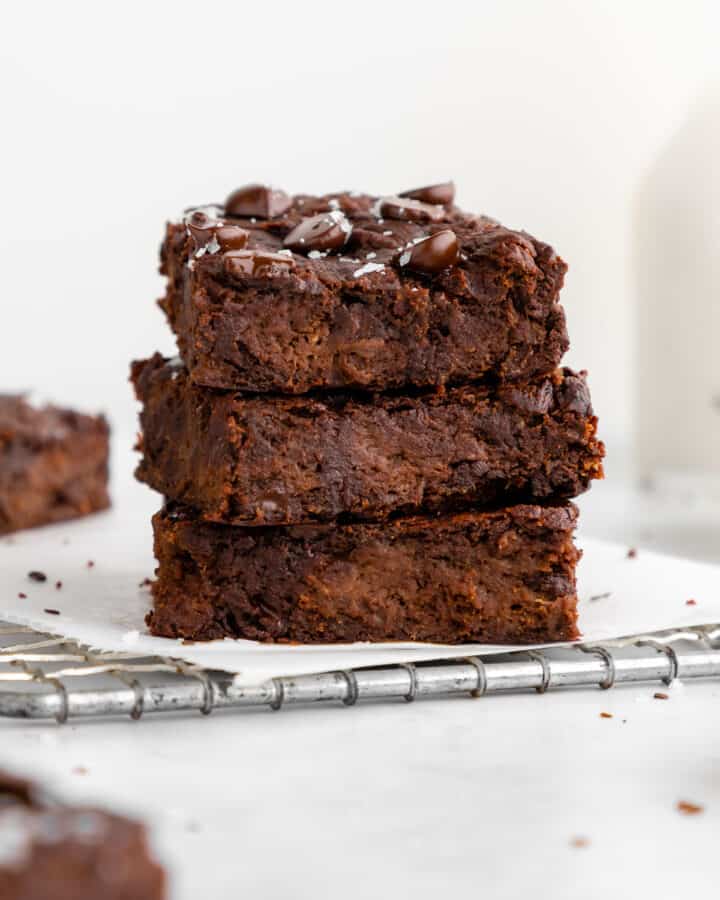 a stack of four flourless vegan protein brownies in front of a bowl of chocolate chips and a bowl of chocolate protein powder