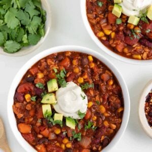 two bowls filled with vegan tempeh chili beside a bamboo fork and bowls of cilantro, corn, and cashew sour cream