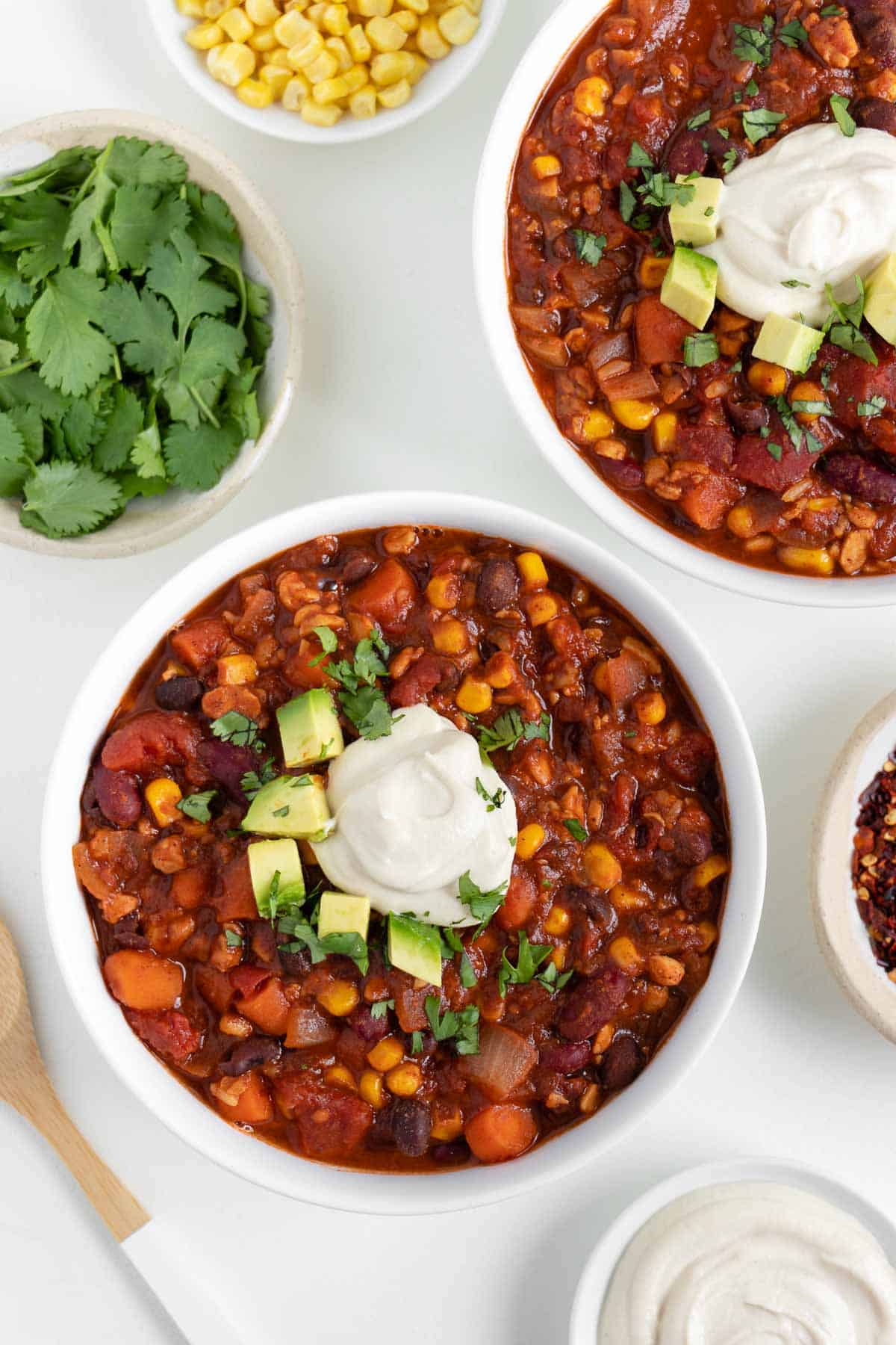 two bowls filled with vegan tempeh chili beside a bamboo fork and bowls of cilantro, corn, and cashew sour cream