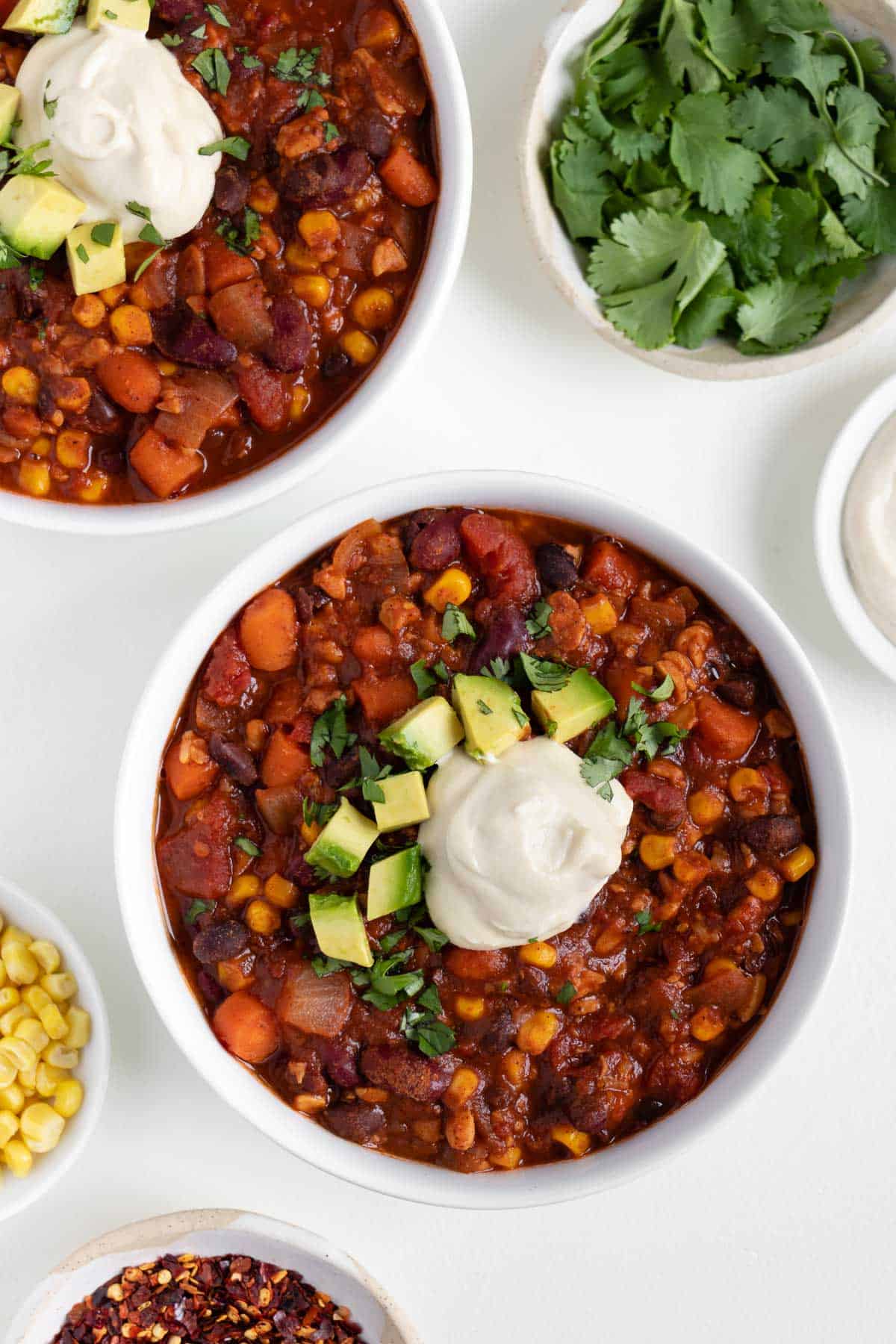 two bowls of vegan tempeh chili surrounded by bowls of cilantro, corn, and cashew sour cream