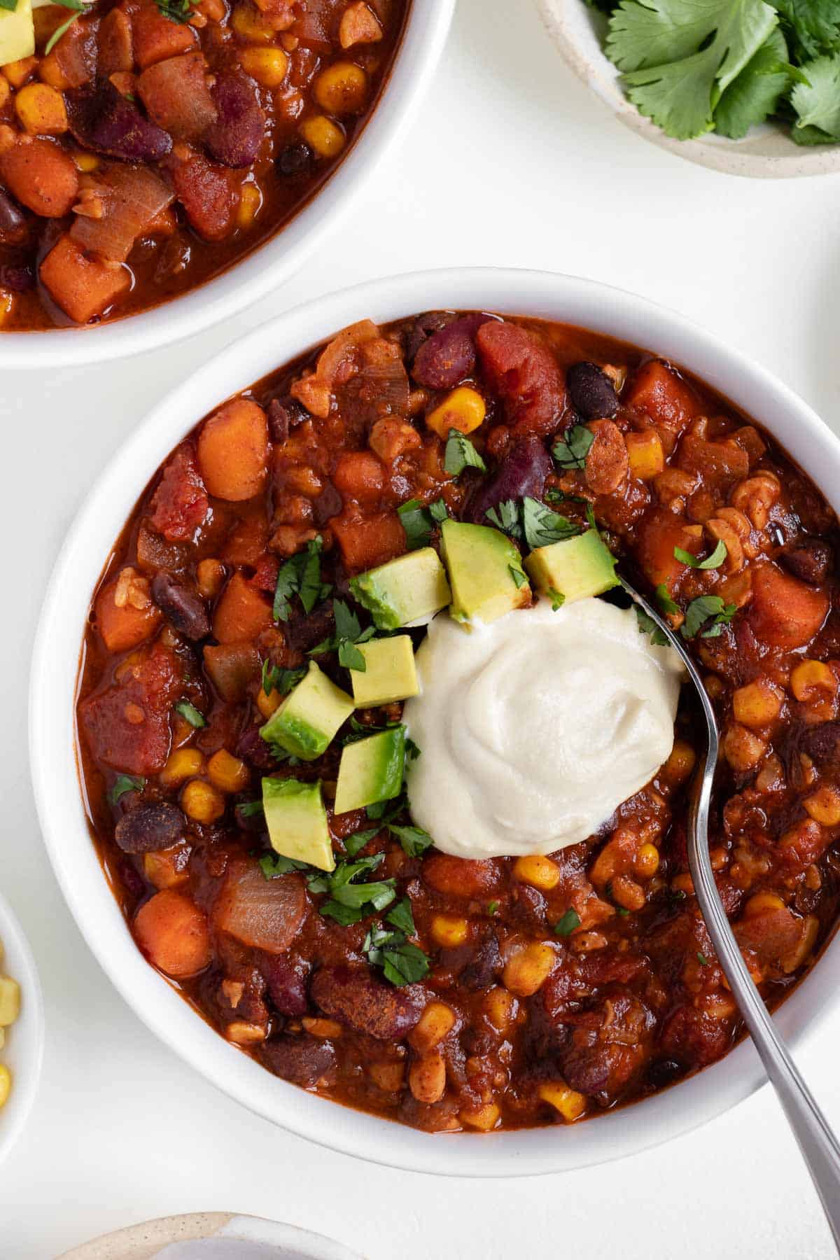 a metal spoon scooping into a bowl of vegan tempeh chili with sour cream, cilantro, and avocado