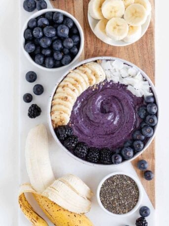 a wood and marble cutting board topped with a blueberry banana smoothie bowl, ripe banana, chia seeds, and berries