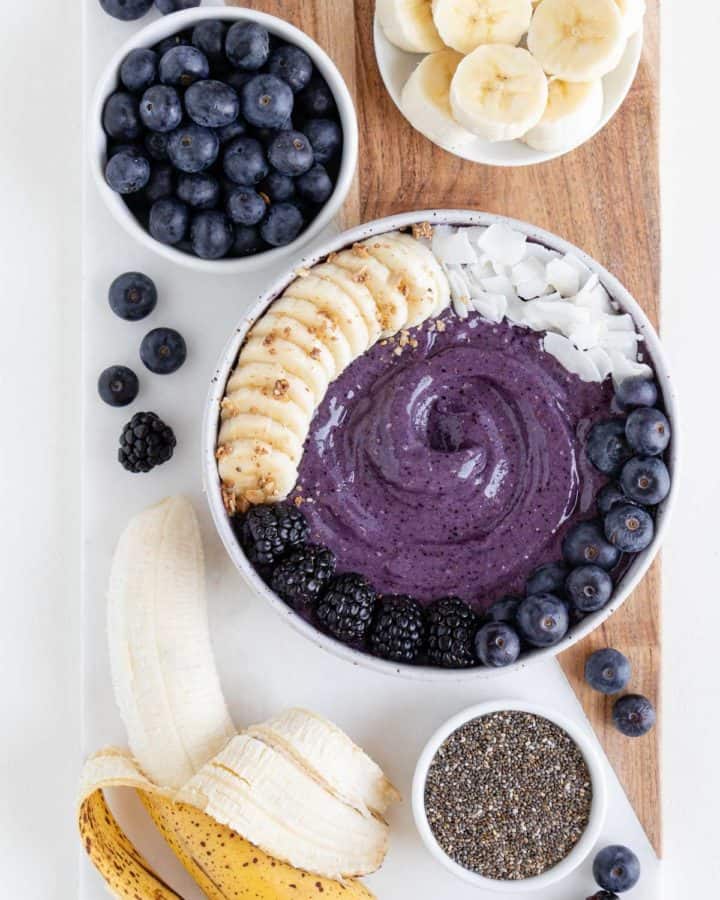a wood and marble cutting board topped with a blueberry banana smoothie bowl, ripe banana, chia seeds, and berries