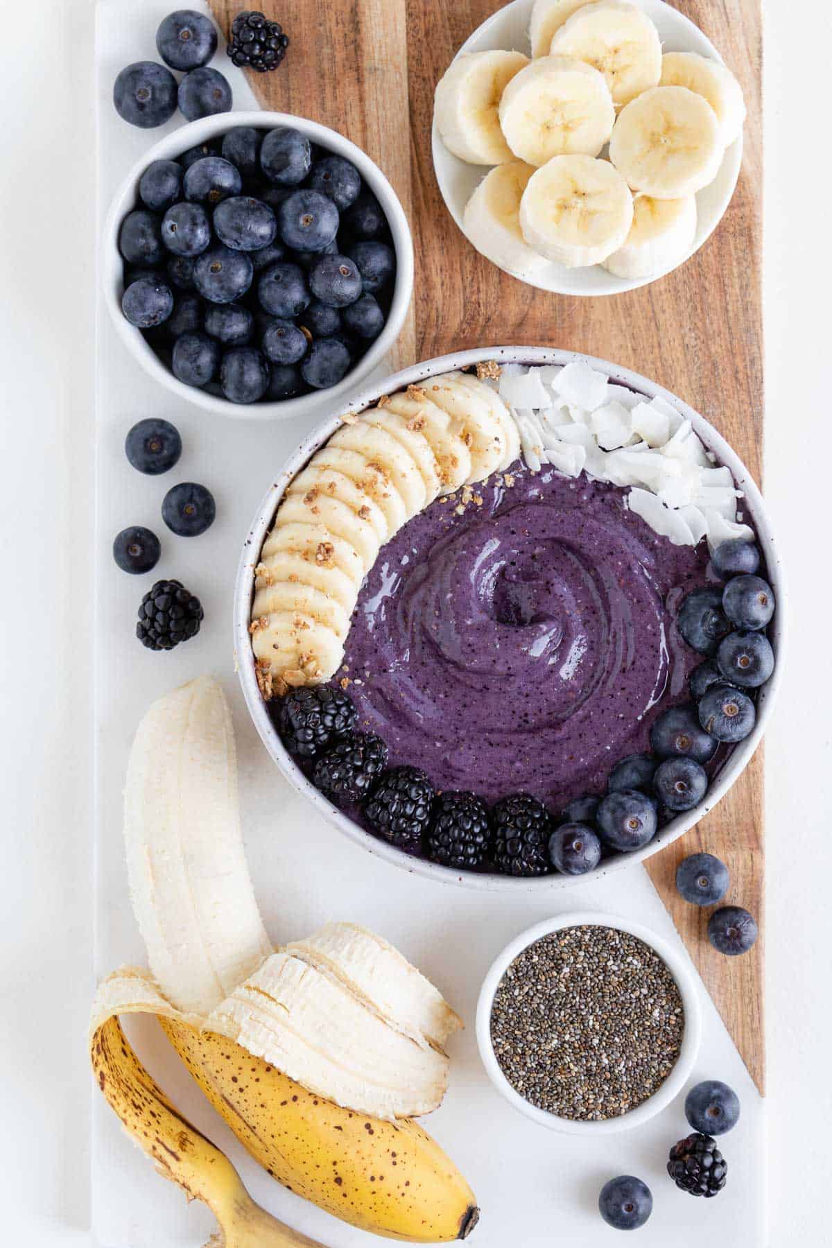 a wood and marble cutting board topped with a blueberry banana smoothie bowl, ripe banana, chia seeds, and berries