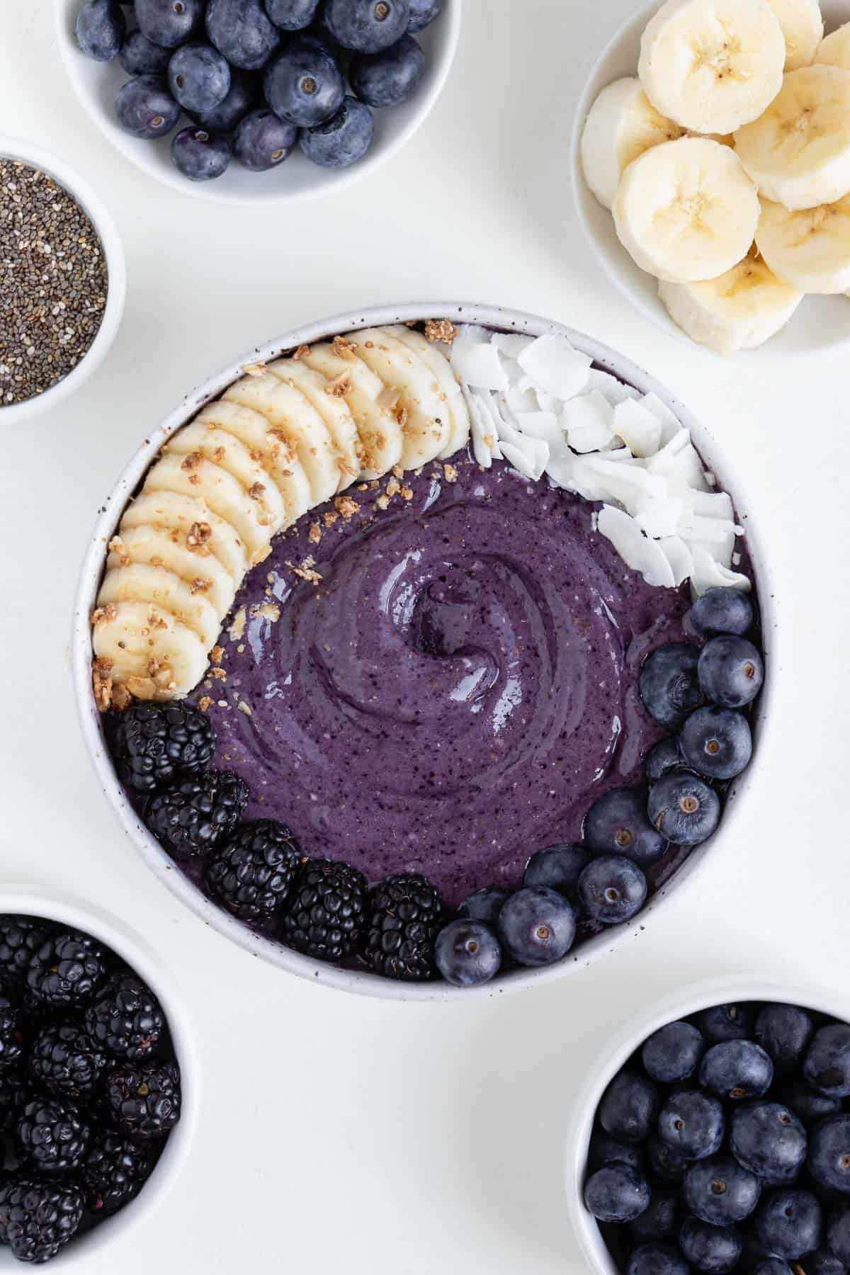 a purple blueberry banana smoothie bowl surrounded by bowls of chia seeds, blueberries, blackberries, and sliced banana
