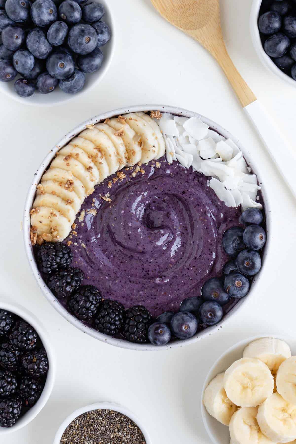 a blueberry banana smoothie bowl surrounded by a wooden spoon, a bowl of berries, a bowl of chia seeds, and a bowl of sliced bananas