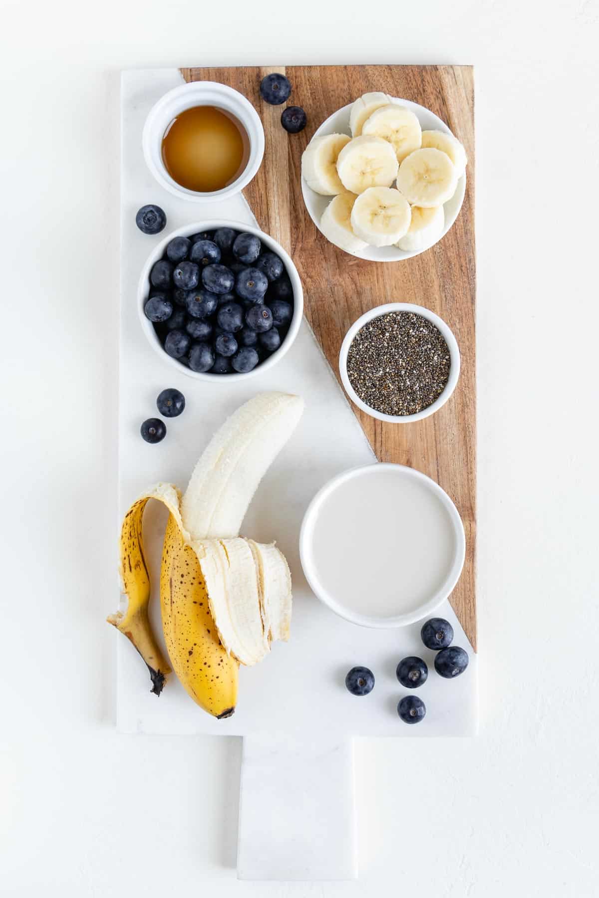 a ripe banana, a bowl of blueberries, a bowl of chia seeds, a glass of almond milk, and a bowl of sliced bananas on a marble cutting board