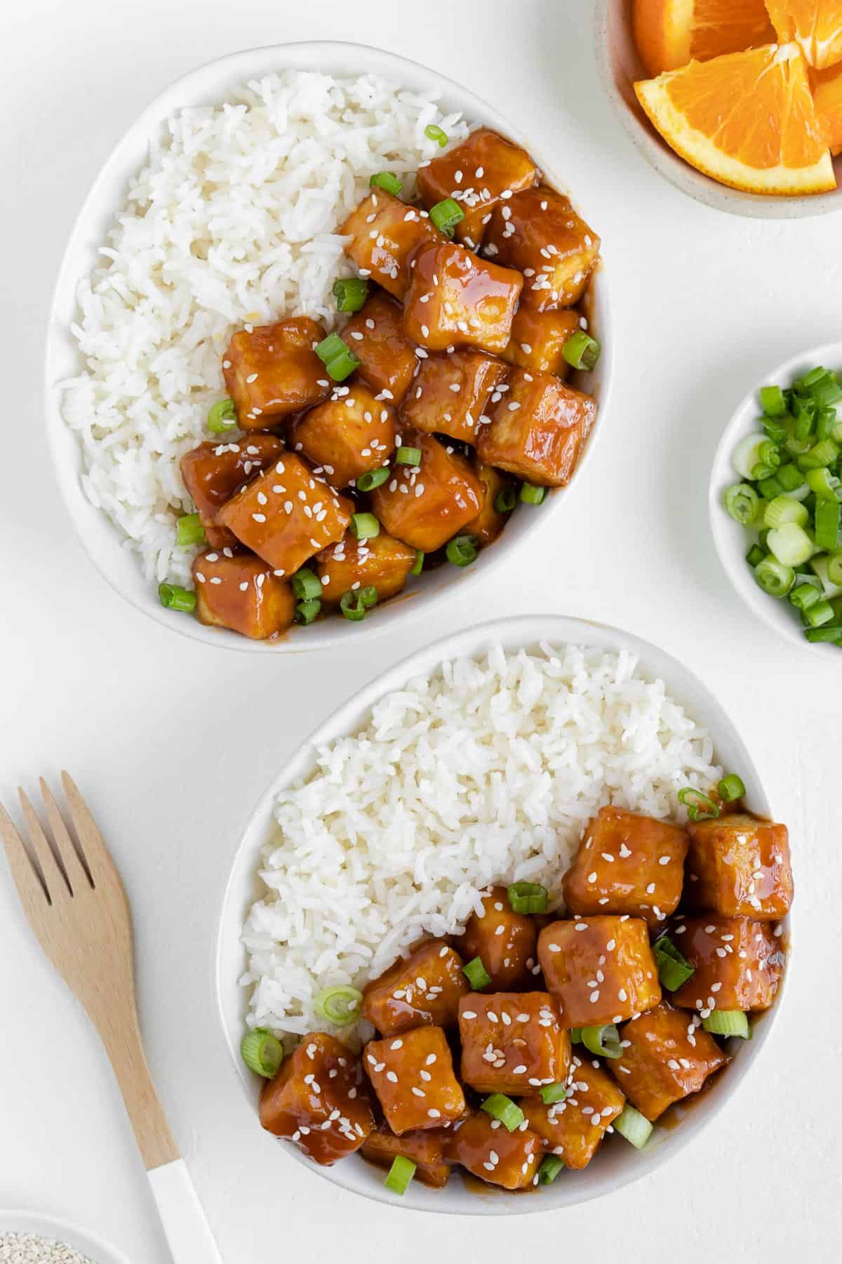 two bowls filled with vegan chinese food beside a bowl of green onions and a wooden fork