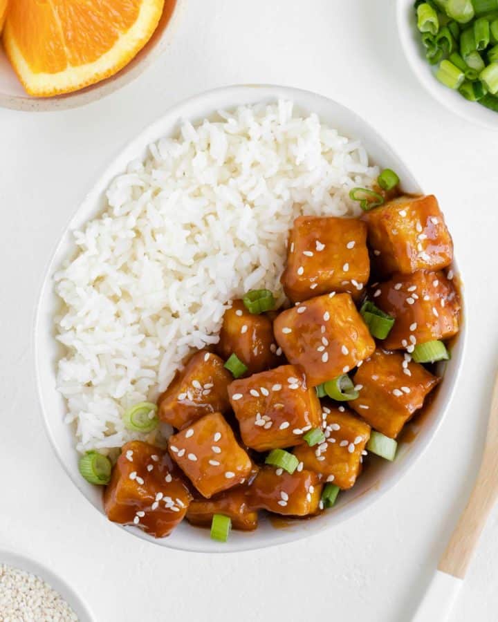 orange tofu in a bowl with white rice, surrounded by a plate of oranges, green onions, and sesame seeds