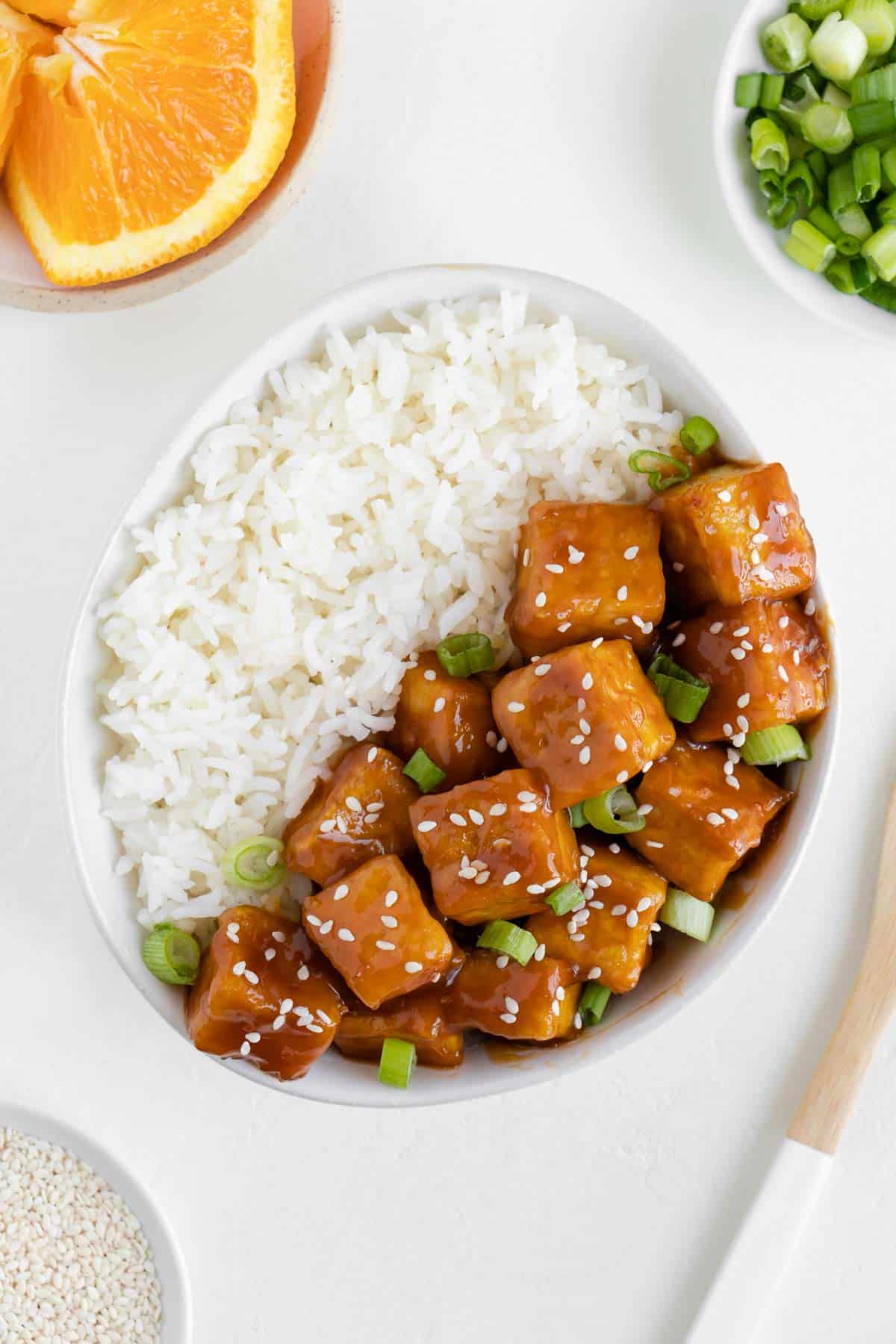 orange tofu in a bowl with white rice, surrounded by a plate of oranges, green onions, and sesame seeds