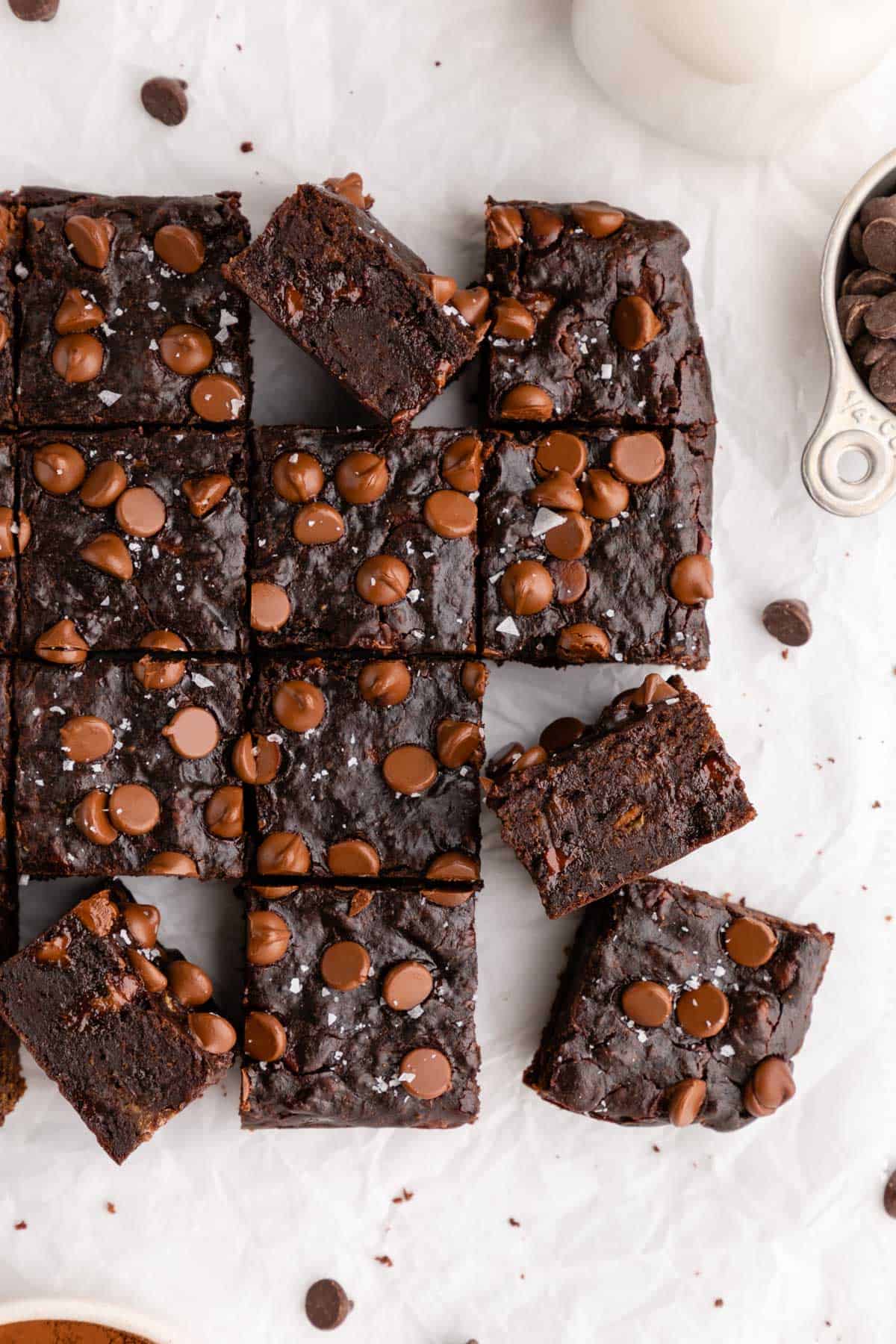 four vegan banana brownies on a white plate surrounded by a glass of almond milk, a bowl of almond butter, a bowl of cocoa powder, and a wooden fork