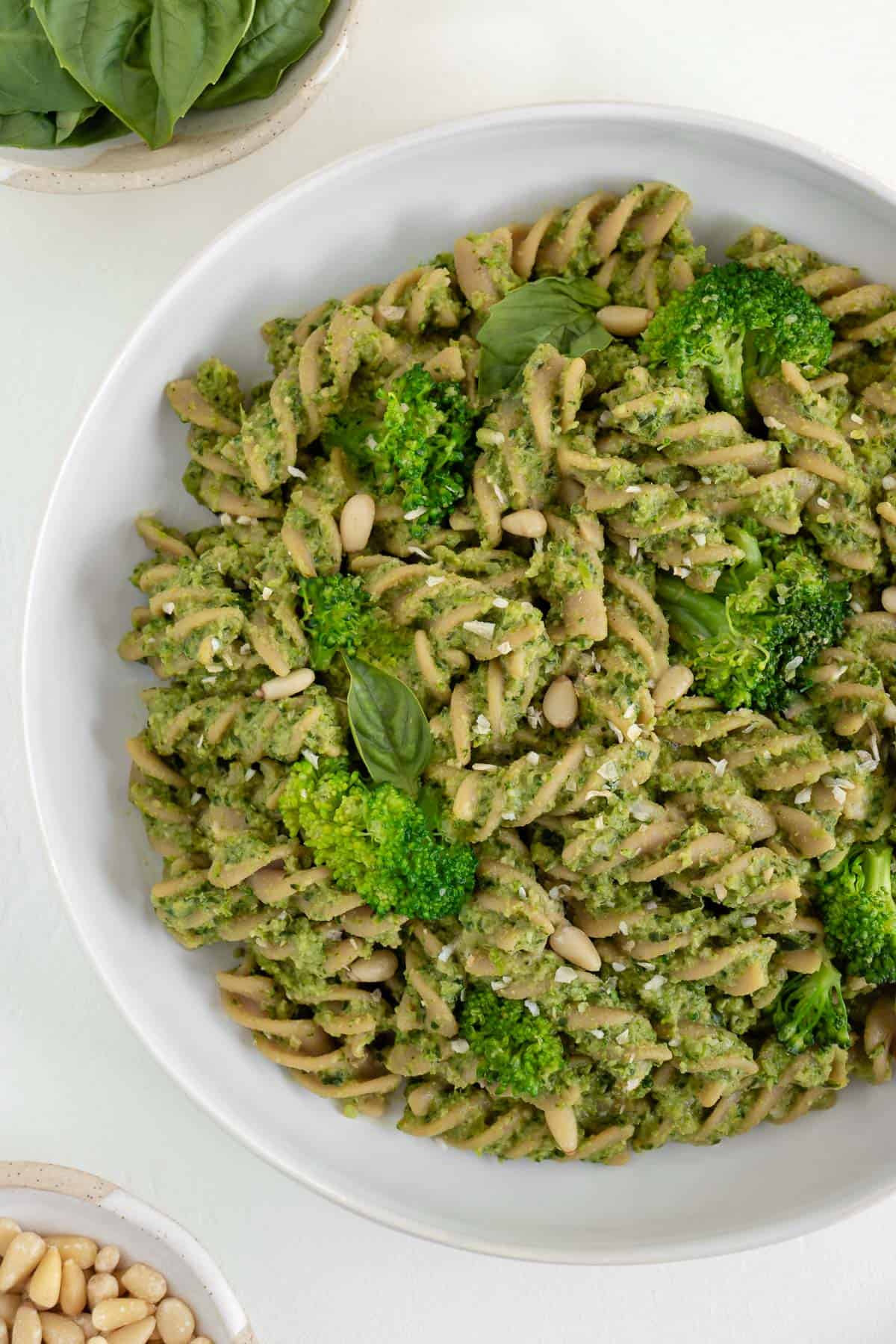 vegan broccoli pesto pasta in a white bowl beside a small bowl filled with basil and another small bowl filled with toasted pine nuts