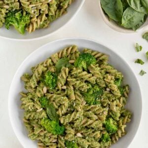 two bowls of vegan broccoli pesto pasta surrounded by a bowl of basil and a bowl of toasted pine nuts